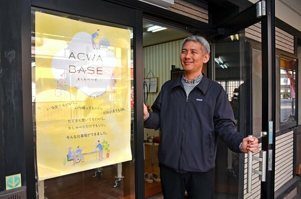 アクワベースの玄関ドアを開ける齊藤社長。ドアには「いつ来てもいいし、いつ帰ってもいい。だれが来てもＯＫ」と記したポスターが張られている