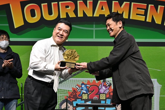 リアルイベント「ウイコレ FAN TOURNAMENT」で連覇を達成し、有田哲平（右）から優勝トロフィーを受け取る山崎弘也