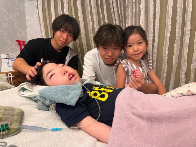 ベッドに横たわる三男の弘一郎くん、ベッドサイド左から順一朗さん、宗一郎さん、末っ子の妹。弘一郎くんは24時間人工呼吸器の管理が必要です。外出時は一大事になることから同じような境遇の人々にボランティア演奏をしようと思いつきました（画像提供：兄―ズ）
