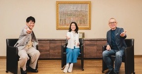 3人の人物が椅子に座り、手を前に差し出している様子。背景には額装された絵画が飾られている。