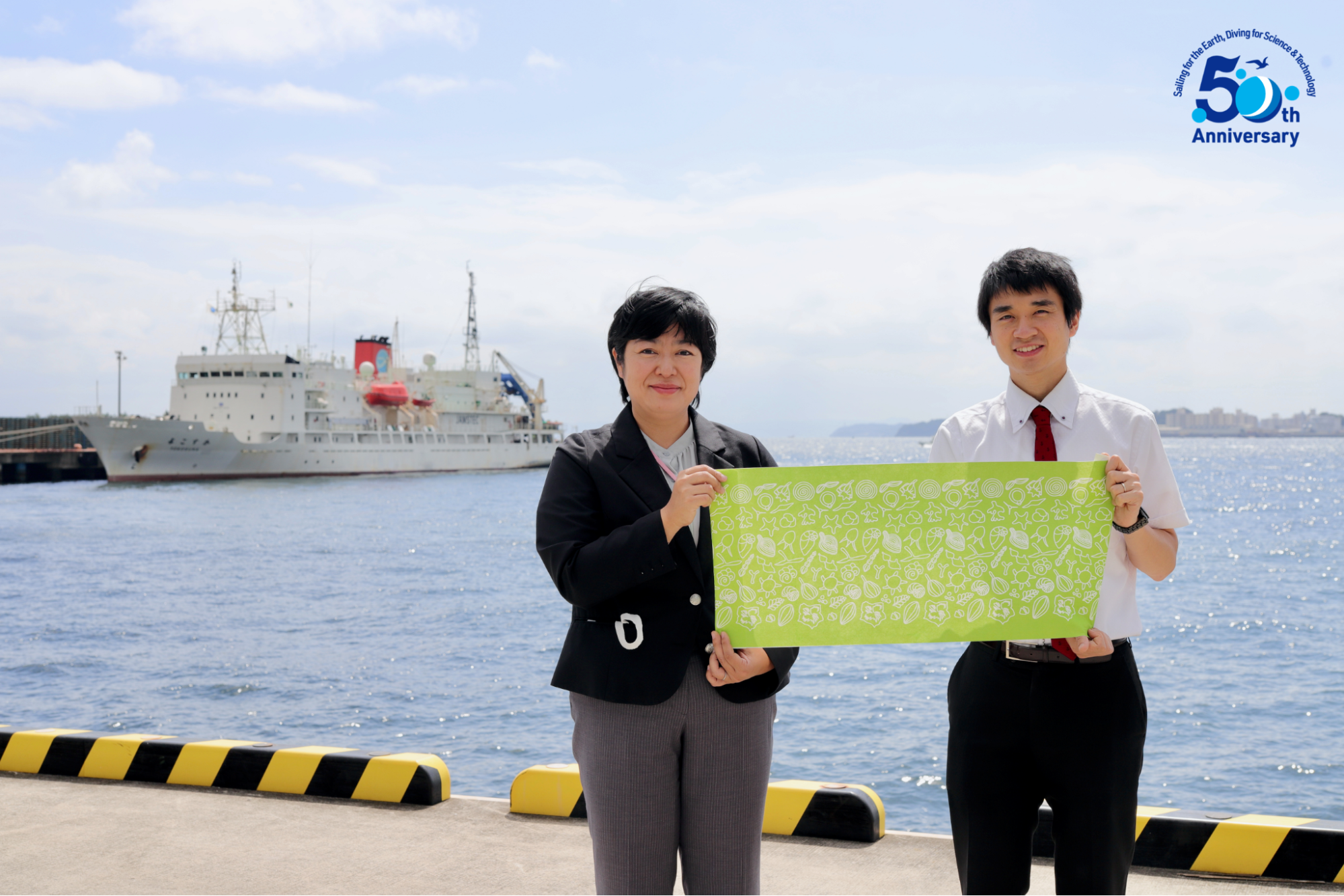 50周年記念切手チームの後藤さん（左）、笠原さん（右）
後藤さんが書いたこの手ぬぐいのモチーフが、よ〜く見ると実は切手シートのあるところに採用されている（写真提供：JAMSTEC）