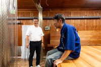 巨大なはかりに腰かけ、田中社長（左）から話を伺う下柳さん
