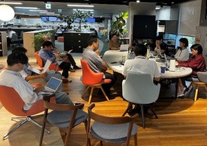 オフィス内のラウンジスペースで、円形テーブルを囲みながら複数の人が打ち合わせをしている様子。周囲では椅子に座って資料やパソコンを見る人もいる。