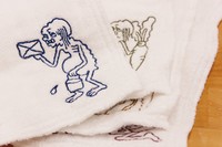 「拭き掃除をする餓鬼」など、白地のふきん右下部分にワンポイント、キャラクターが刺繍されています（提供写真）