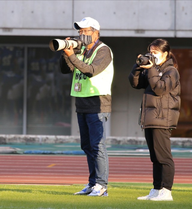 若い記者とともにアメリカンフットボールの試合を撮影する山岡丈士さん（左）＝2021年11月、ヤンマースタジアム長居