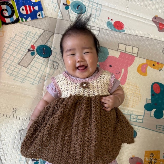 お向かいのおばあちゃんが編んだワンピースを着て、笑顔を見せる生後6カ月の娘／「あーちゃんちゃん」さん（@aydayo221）提供