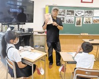 漫画を手に「表紙の猫２匹は何を運んでいますか」と児童に質問する三田さん＝７月１日（画像の一部を加工しています）