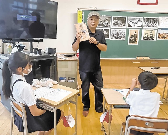 漫画を手に「表紙の猫２匹は何を運んでいますか」と児童に質問する三田さん＝７月１日（画像の一部を加工しています）