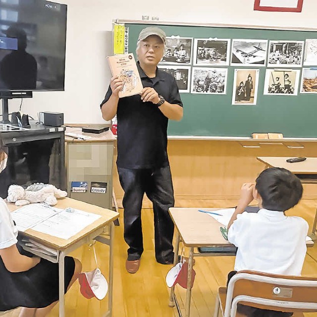 漫画を手に「表紙の猫２匹は何を運んでいますか」と児童に質問する三田さん＝７月１日（画像の一部を加工しています）