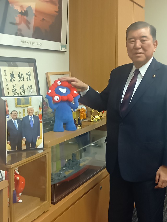 トランプ米大統領との写真を前に、「ミャクミャク」の人形に手を置く石破茂前首相＝東京・永田町の衆議院第二議員会館